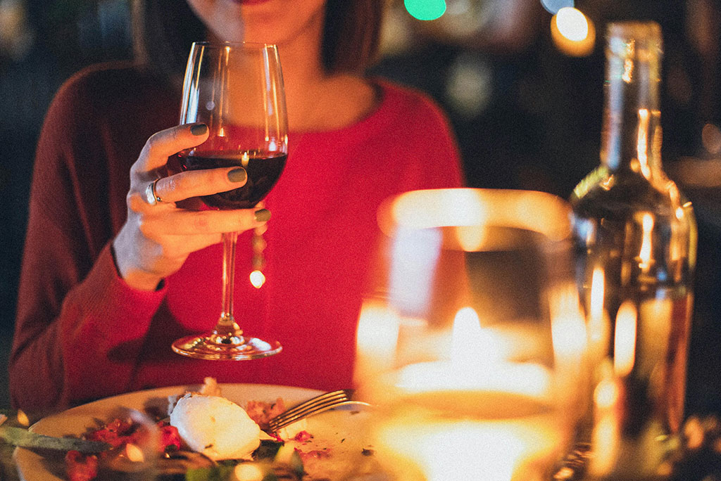 a woman sitting at a set table drinking a glass of red wine